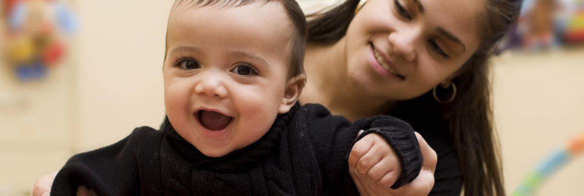 picture of smiling baby being propped up from behind by a young woman with long brown hair looking over the baby's left shoulder