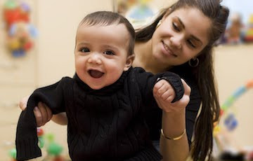 picture of smiling baby being propped up from behind by a young woman with long brown hair looking over the baby's left shoulder